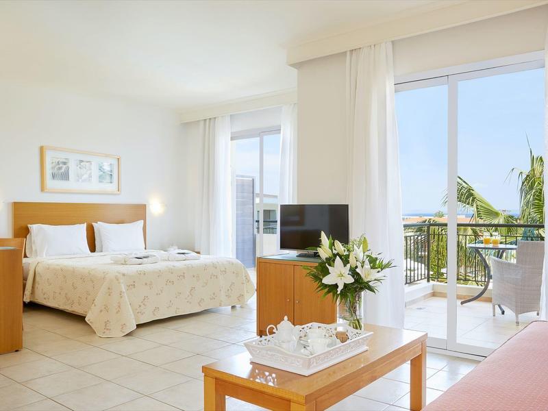 Jasny pokój hotelowy z podwójnym łóżkiem, drewnianymi meblami i balkonem z widokiem na morze.