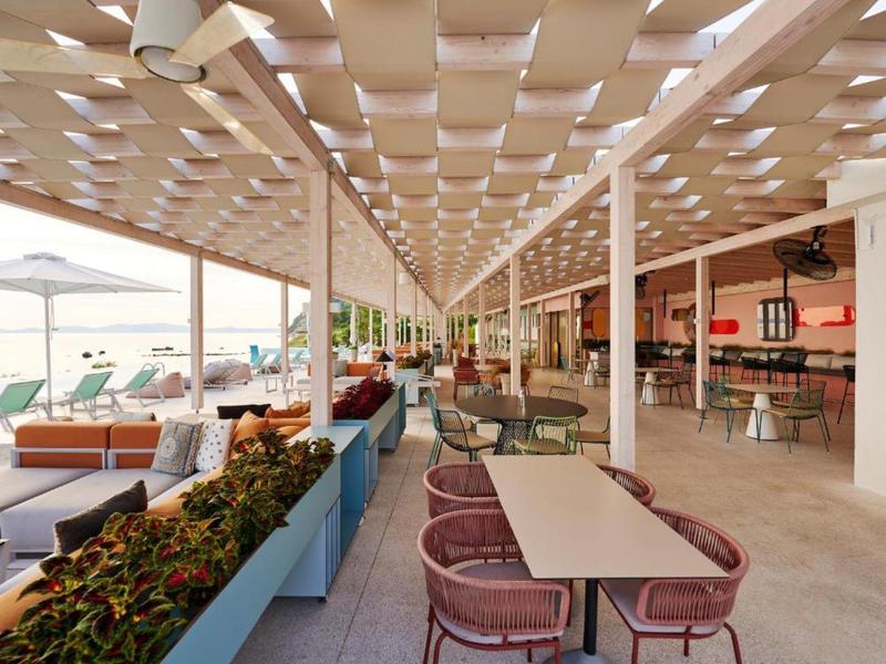 Modern hotel terrace with tables, chairs, umbrellas, and a view of the swimming pool.