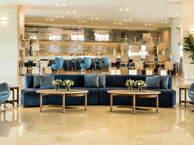 Area lounge moderna in hotel con poltrone blu e tavolini dorati davanti al bancone del bar.