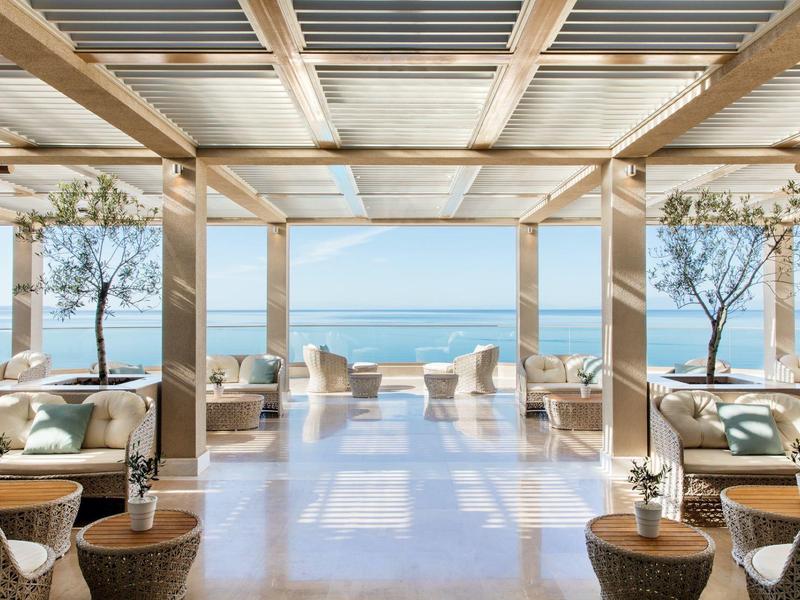 Area lounge moderna e aperta con vista sul mare e terrazza, arredi chiari e piante.