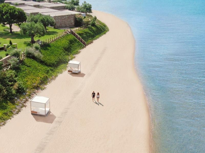 Zwei Personen spazieren an einem breiten Strand mit Strandkörben und klarem Wasser.