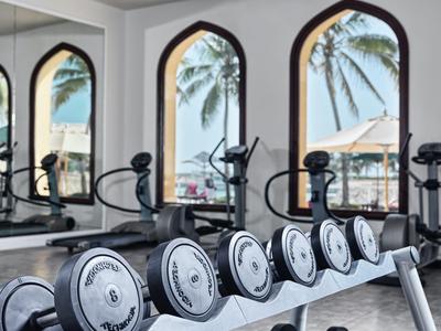 Gym with dumbbells in front of windows showing sea view and palm trees.