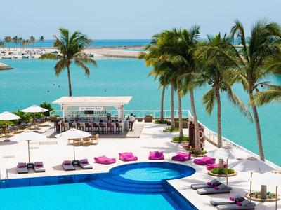 Moderní bazénová oblast s lehátky, palmami a výhledem na tyrkysovou vodu a marinu.