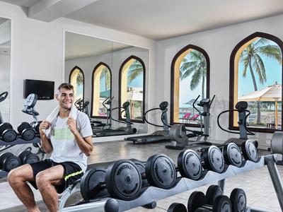 Man sitting in a hotel gym holding a water bottle near dumbbells and exercise machines.