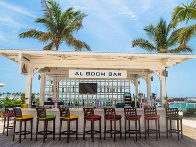 Otevřený plážový bar s barevnými stoličkami a palmami v pozadí pod modrou oblohou.