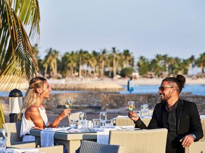Pár si užívá víno u venkovního stolu s palmami a pláží v pozadí.