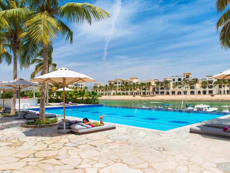 Velká bazénová plocha s lehátky a slunečníky před hotelem pod modrou oblohou.