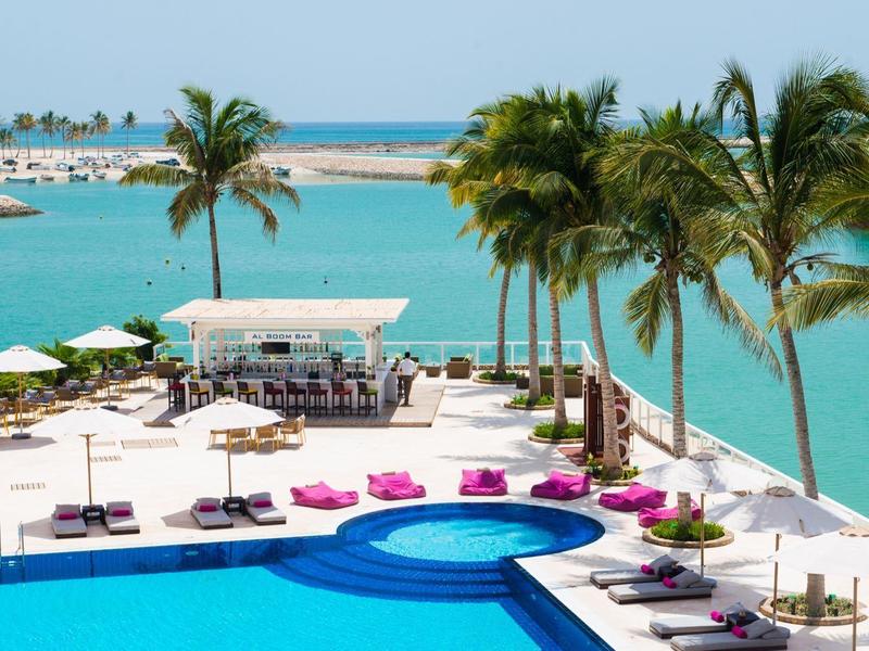 Moderní bazénová oblast s lehátky, palmami a výhledem na tyrkysovou vodu a marinu.