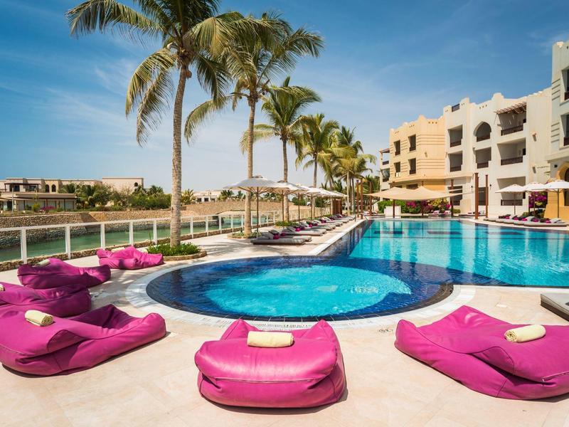 Moderní hotelový komplex s bazénem, palmami a fialovými lehátky pod modrou oblohou.