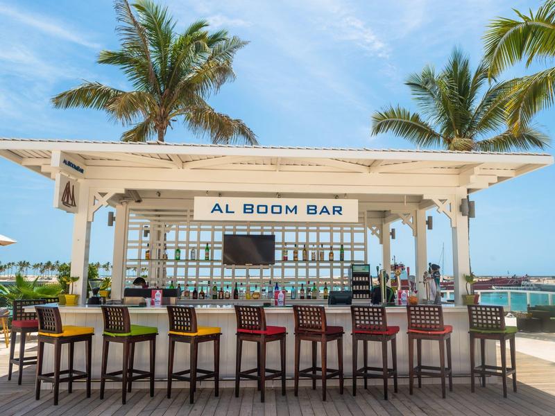 Otevřený plážový bar s barevnými stoličkami a palmami v pozadí pod modrou oblohou.