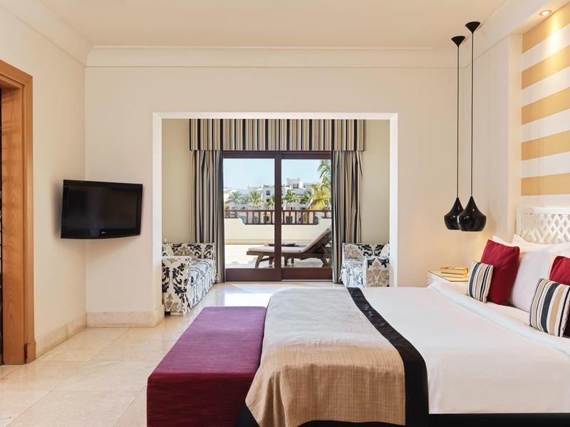 Světý hotelový pokoj s manželskou postelí, TV a výhledem na terasu s lehátky.