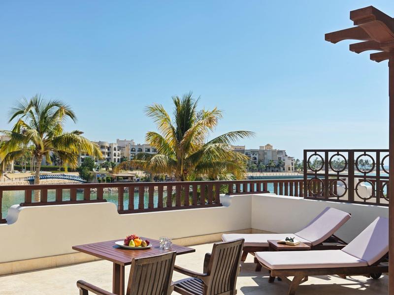 Terasa se dvěma lehátky, stolem a palmami s výhledem na budovy za jasné oblohy.