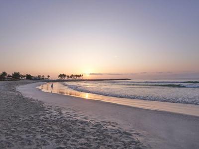 Sandiger Strand mit ruhigem Meer und Sonnenuntergang am Horizont, Palmen im Hintergrund.