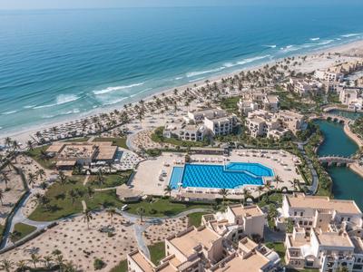 Kurort mit großem blauen Pool, Sandstrand, Palmen und Meer am Horizont unter blauem Himmel.