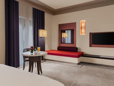 Moderner Hotelraum mit rotem Sofa, rundem Tisch, Stühlen und großen Fenstern mit Vorhängen.