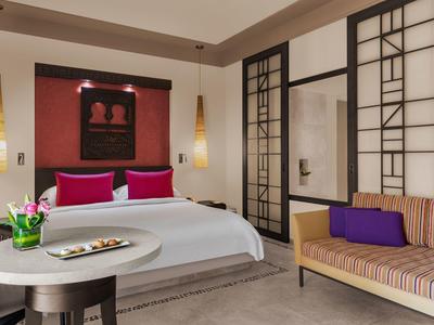 Modernes Schlafzimmer mit großem Bett, rosa Kissen, seitlichen Lampen und Sofa mit lila Kissen.