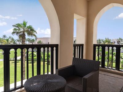 Balkon mit schwarzem Korbsessel, Tisch und Blick auf grüne Wiese und Palmen unter blauem Himmel.