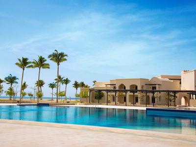 Ein großer Hotelpool mit Palmen und einem Gebäude im Hintergrund unter blauem Himmel.