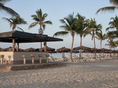 Strandbar mit weißen Hockern und Sonnenschirmen, umgeben von Palmen bei klarem Himmel.