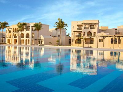 Ein ruhiger Pool mit reflexivem Wasser vor mediterranen Gebäuden und Palmen bei Sonnenuntergang.