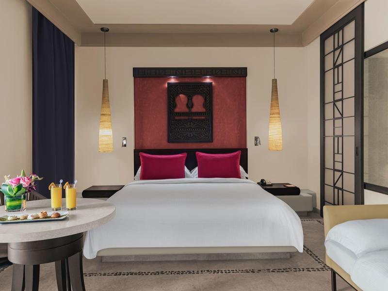 Modernes Schlafzimmer mit weißem Bett, roten Kissen, hängenden Lampen und einem runden Tisch mit Getränken.