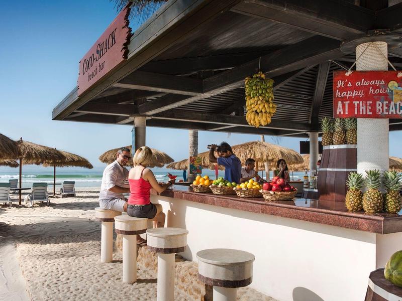 Strandbar mit Holzhockern, frischem Obst und Gästen, Palmen und Meer im Hintergrund bei sonnigem Wetter.