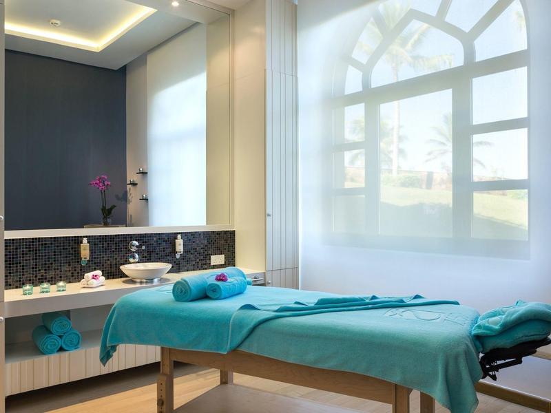 Helles Behandlungszimmer mit massiertisch, blauer Decke, großen Fenstern und moderner Beleuchtung.