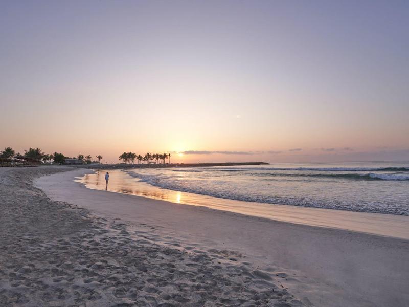 Sandiger Strand mit ruhigem Meer und Sonnenuntergang am Horizont, Palmen im Hintergrund.