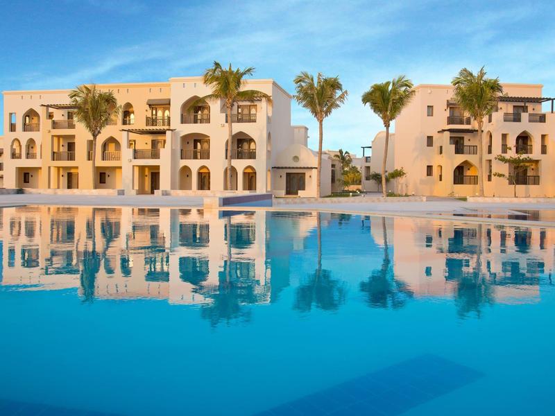 Großer blauer Pool vor zwei hellen Wohngebäuden mit Palmen an sonnigem Himmel.