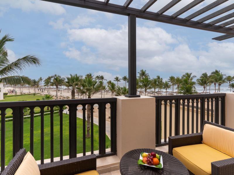Balkon mit gelbem Sofa, Obstteller und Holzgeländer, Blick auf Palmen und blauen Himmel.