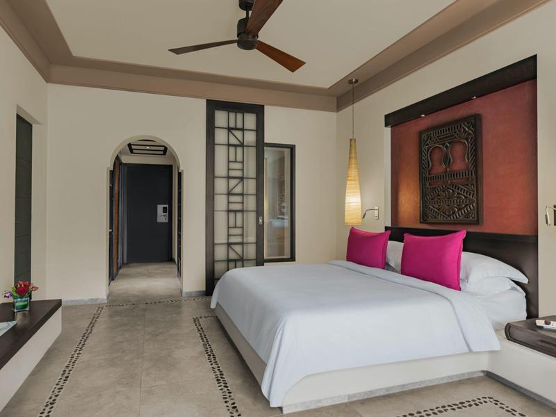 Schlafzimmer mit großem Bett, rosa Kissen, dunklem Wandbild, zwei Hängeleuchten und Ventilator an der Decke.