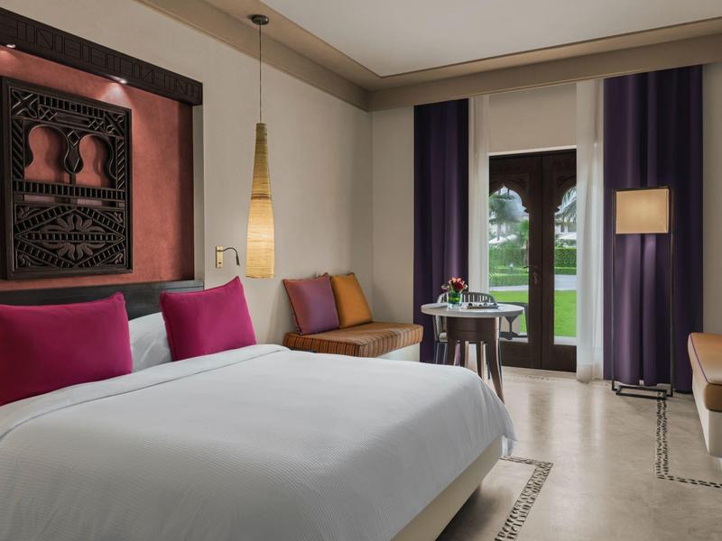 Modernes Schlafzimmer mit großem Bett, bunten Kissen, Fenster mit Vorhängen und gemütlichen Sitzbereichen.