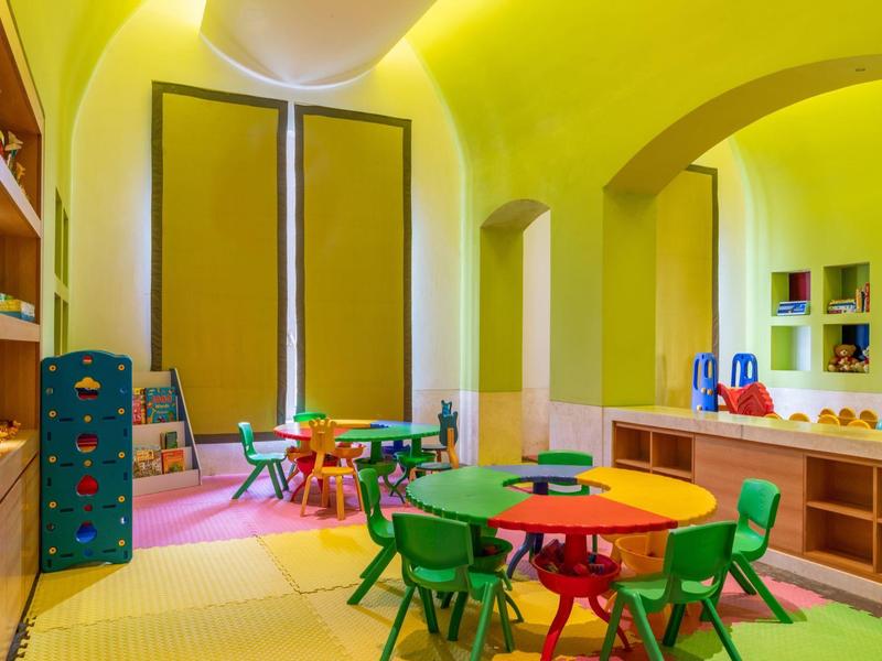 Bunter Kindergartenraum mit Tischen, Stühlen und großen Fenstern, gelbe Wände, verspielte Atmosphäre.