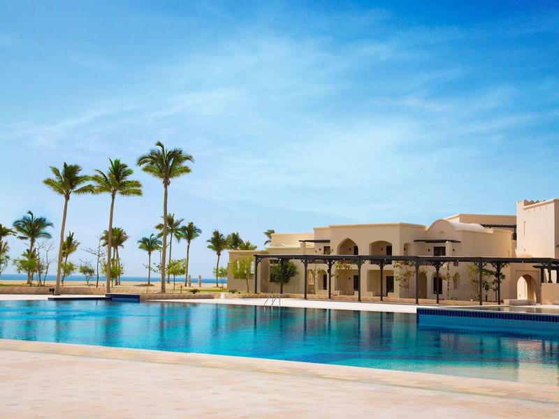 Großer Pool mit blauer Wasseroberfläche, Palmen und modernem beigefarbenem Gebäude bei klarem Himmel.