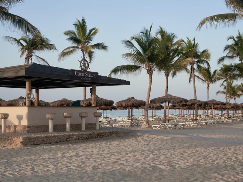 Strandbar mit weißen Hockern und Sonnenschirmen, umgeben von Palmen bei klarem Himmel.