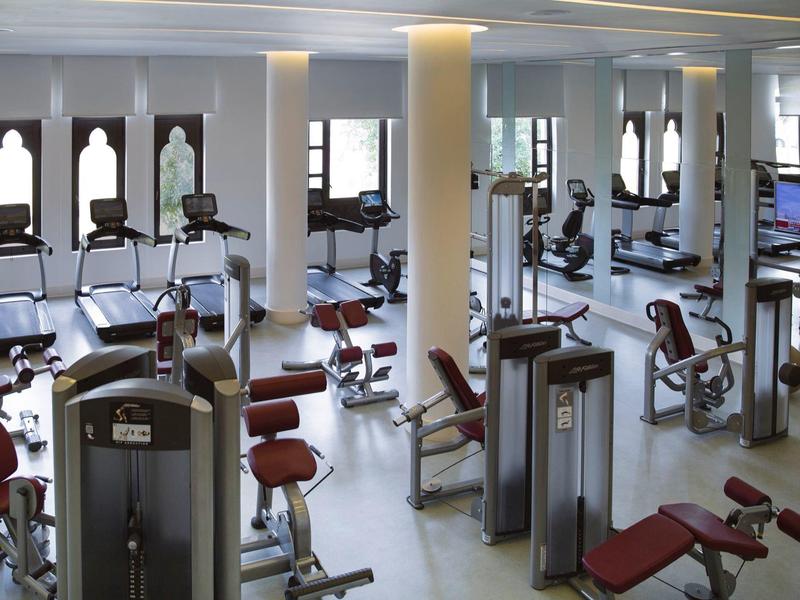 Heller Fitnessraum mit Kraftgeräten, Spiegelwänden und roten gepolsterten Sitzen.