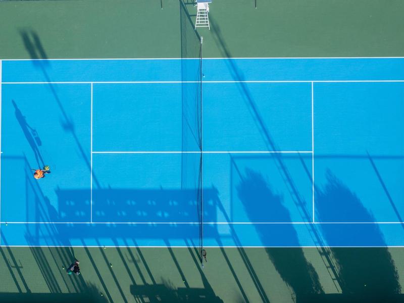 Vogelperspektive auf blauen Tennisplatz mit Spieler, Netzschatten und langen Schatten.