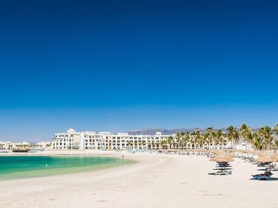 Breiter Sandstrand mit Liegestühlen, Palmen und weißer Hotelanlage unter klarem blauem Himmel.