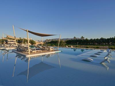 Großer Pool mit Sonnensegel und Liegen, umgeben von Palmen und klarem blauem Himmel.