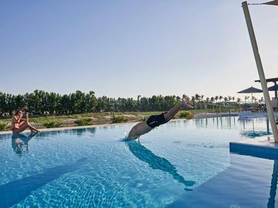 Mann taucht kopfüber in großen blauen Pool, Frau sitzt am Beckenrand bei sonnigem Himmel.