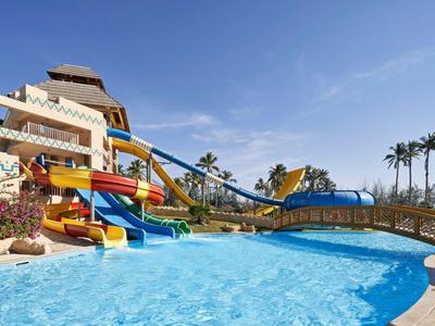 Wasserpark mit bunten Rutschen, großem Pool, Palmen und klarem blauem Himmel im Hintergrund.