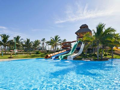 Großer Swimmingpool mit Wasserrutschen und Palmen unter blauem Himmel in tropischer Anlage.