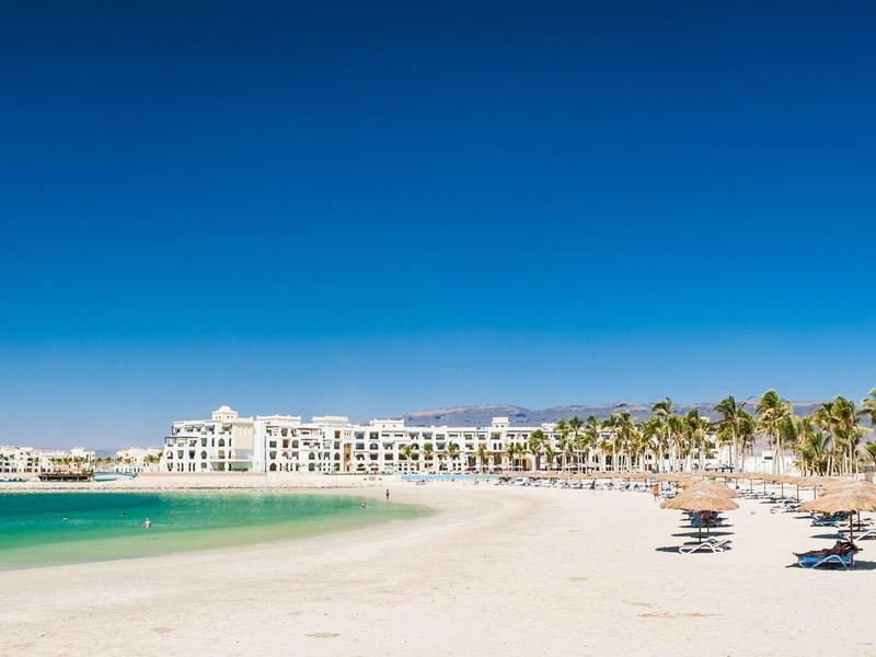 Breiter Sandstrand mit Liegestühlen, Palmen und weißer Hotelanlage unter klarem blauem Himmel.