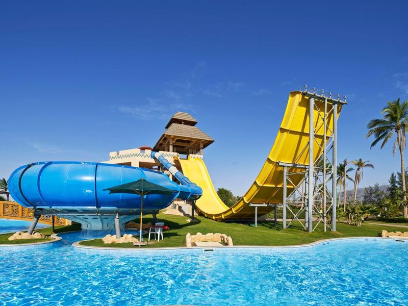 Wasserpark mit blauem Pool, großen gelben und blauen Wasserrutschen und Palmen im Hintergrund.
