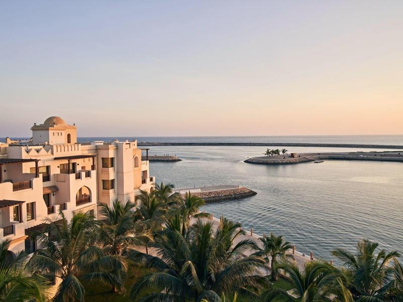 Meerblick mit Hafen, Palmen und weißen Gebäuden bei Sonnenuntergang.