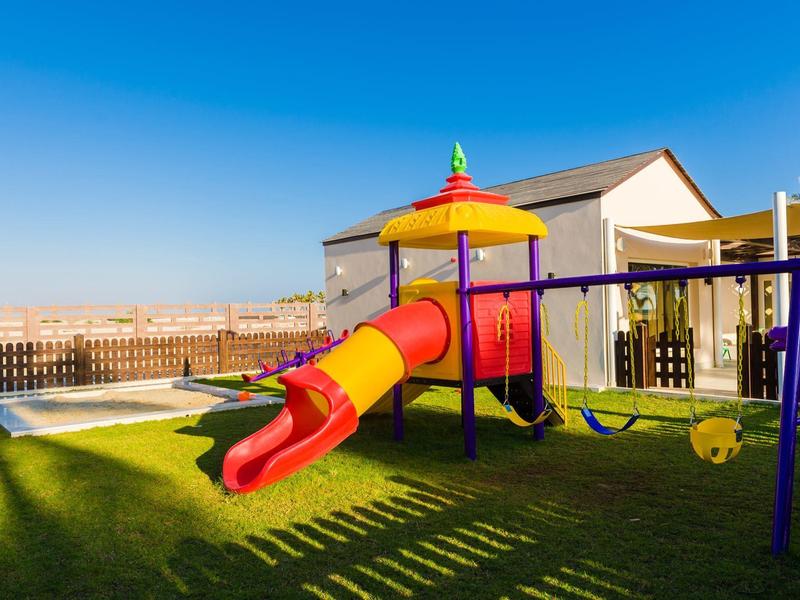 Bunter Spielplatz mit Rutsche, Schaukel und Klettergerüst auf grünem Rasen unter blauem Himmel.