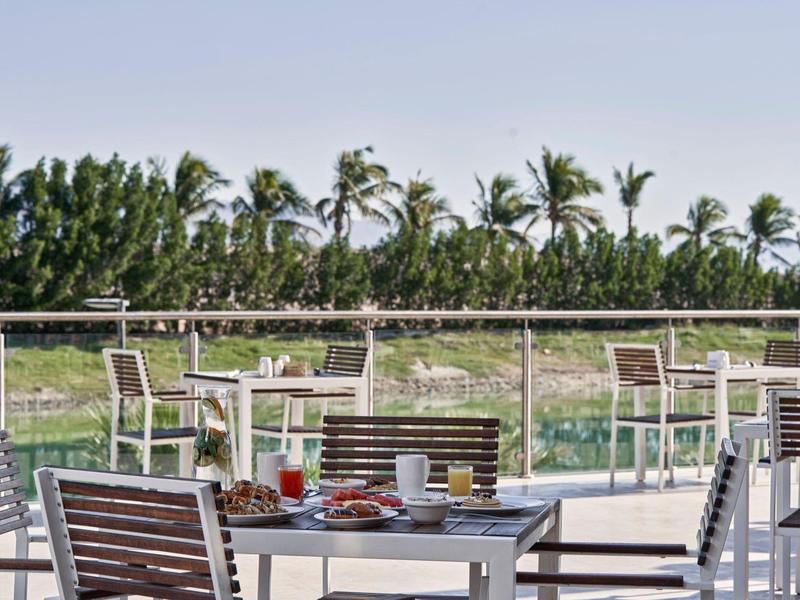 Terrassenbereich mit Tischen, Stühlen und Essen, darunter grüne Palmen und blauer Himmel.