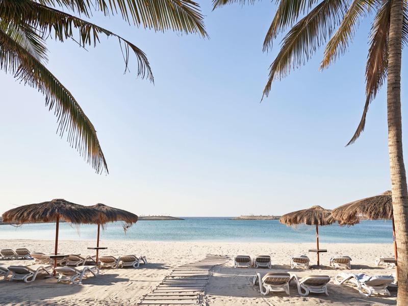 Sandstrand mit Liegestühlen und Palmen, weiter Blick auf ruhiges blaues Meer unter klarem Himmel.