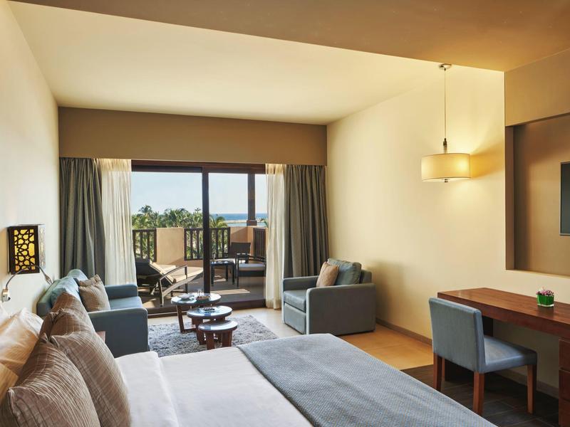 Helles Hotelzimmer mit Doppelbett, Sofa, Schreibtisch und großem Fenster zur Terrasse mit Blick ins Freie.