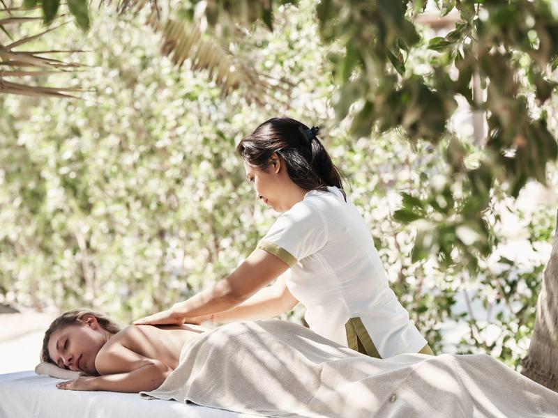 Frau bekommt Rückenmassage auf Massagetisch im Garten unter grünen Blättern.